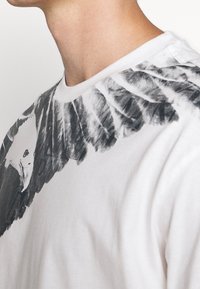 Bílý bavlněný tričko s černobílou grafikou peří a hlavy ptáka na rameni, kulatý výstřih, krátké rukávy.