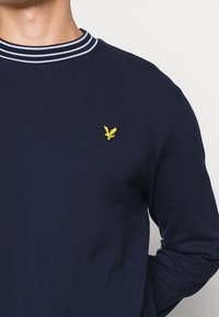 Felpa blu navy con colletto a girocollo a coste, caratterizzata da strisce bianche. Logo di un'aquila gialla ricamato sul lato sinistro del petto. Tessuto morbido e testurizzato.