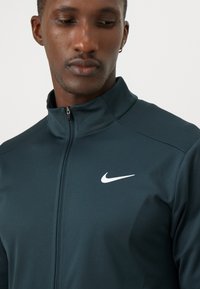 Nike'i tumedast külast valmistatud jope, millel on kõrge krae, lukuga closure ja valge logo rinnal. Sile, tekstureeritud kangas.