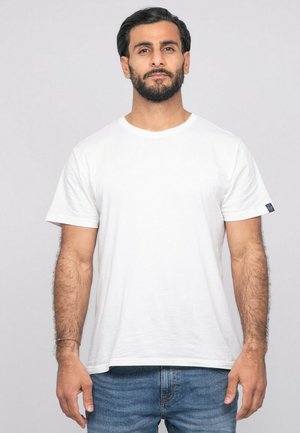 Homme aux cheveux foncés et à la barbe, portant un t-shirt blanc uni à manches courtes et un jean bleu, debout devant un fond neutre.