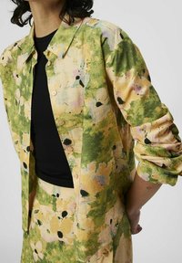 Chemise à boutons florale jaune et verte en tissu texturé léger ; elle présente une coupe décontractée et des manches retroussées.