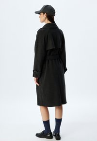 Donkergrijze wollen jas met een broekriem in de taille, drop shoulder ontwerp en knoopaccenten bij de manchetten, gecombineerd met een grijze pet, marineblauwe sokken en zwarte schoenen.