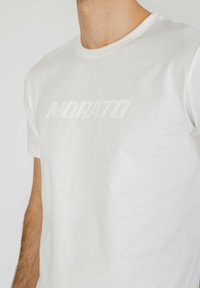 Camiseta de algodón blanca con cuello redondo, que presenta un texto "MORATO" en relieve y brillo en el pecho. Textura suave y diseño minimalista.