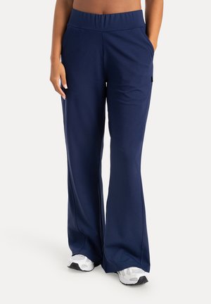 Persona con pantalones anchos azul marino y zapatillas blancas, de pie con una mano en el bolsillo contra un fondo liso.