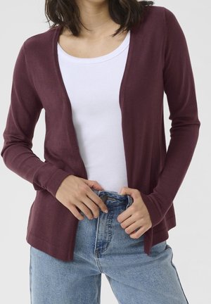 Frau trägt ein weißes geripptes Tanktop, eine offene dunkelviolette Strickjacke und hellblaue Jeans und richtet die Jeans an der Taille.