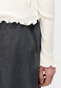 Κοντινό πλάνο ενός ατόμου που φοράει εκρού πλεκτό μπλουζάκι με μακριά μανίκια και κυματιστά άκρα και σκουρόχρωμο τζιν με εμφανείς πορτοκαλί ραφές.