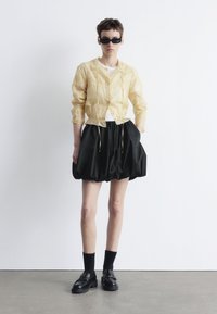 Yellow lace-trimmed jacket with drawstring waist, paired with a voluminous black skirt. Model wears black loafers and sunglasses.