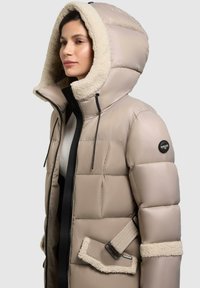 Giubbotto imbottito beige con cappuccio foderato in peluche, chiusura con zip e rifiniture in pile su tasche e polsini. Presenta un logo a patch sulla manica.