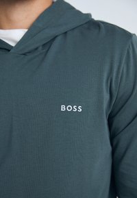 Mikina s kapucňou z tmavozelenej tkaniny s hladkým povrchom. Obsahuje biele vyšívané logo "BOSS" na hrudi.