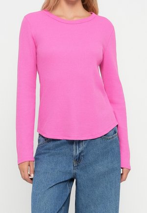 Femme portant un pull côtelé à manches longues de couleur rose vif associé à un jean bleu délavé ample, visible des épaules aux hanches.
