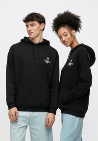 Čierne bavlnené mikiny s kapucňou, prednými vreckami, nastaviteľnými sťahovacími šnúrkami a grafickým dizajnom na hrudi, v kombinácii so svetlomodrými džínsami.