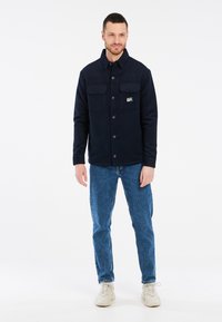 Jūrinės tamsiai mėlynos vilnos striukės su dviem krūtinės kišenėmis ir mygtukais, derinamos su mėlynais džinsais ir šviesių spalvų sportbačiais, stovinčiais ant balto fono.