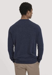 Dunkelblauer Pullover aus strukturiertem Strickstoff. Hat einen Rundhalsausschnitt und gerippte Bündchen, kombiniert mit beigen Hosen. Sicht von hinten.
