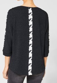 Mörkgrå långärmad tröja med vit och svart hundtandsrand som löper vertikalt nedför ryggen och ärmarna, bärs av en stående person.