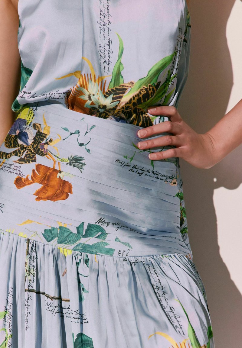 Robe sans manches avec taille plissée, présentant des motifs floraux et feuillus colorés ainsi que des accents d’écriture manuscrite sur un tissu léger.