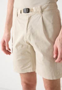 Béžové šortky vyrobené z lehkého materiálu, s plochým předním dílem, černým nastavitelným opaskem a bočními kapsami, s uvolněným střihem.