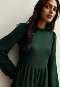Ciemnozielona prążkowana sukienka z okrągłym dekoltem, długimi bufkami i rozkloszowanym dołem. Materiał ma fakturowaną powierzchnię, co dodaje wizualnego zainteresowania.
