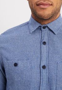 Camicia azzurra con texture, colletto button-down, dotata di bottoni neri e due tasche sul petto. Realizzata in tessuto tessuto con un motivo sottile.