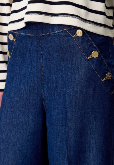 Pantalones de mezclilla azul oscuro con botones dorados en los bolsillos en ángulo, combinados con una blusa de rayas blanca y negra.