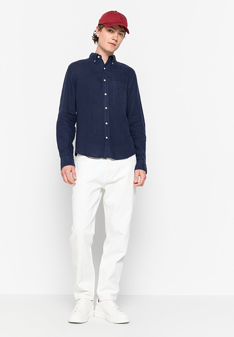 Camisa de botones azul marino con mangas largas, bolsillo en el pecho izquierdo; combinada con pantalones blancos y una gorra roja; sobre un fondo sencillo.