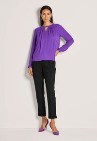 Paarse langemouwen top met geplooide details en een keyhole halslijn, gecombineerd met zwarte cropped broeken en paarse hoge hakken.