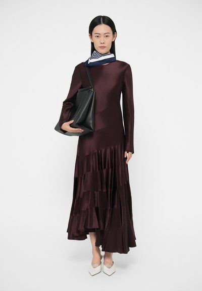 Burgundska satenasta obleka z dolgimi rokavi in večplastnim robom z volani. Model drži črno usnjeno torbico in nosi modro-beli vzorčasti šal.