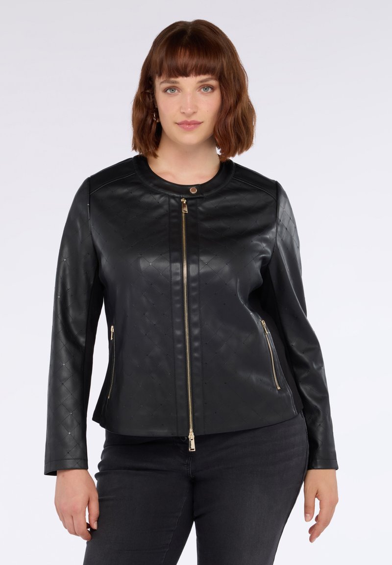 Donna con capelli castani fino alle spalle che indossa una giacca nera in ecopelle trapuntata con zip frontale e tasche laterali con zip, e pantaloni neri.