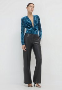 Donna che indossa un top blu in velluto con scollatura a V profonda, pantaloni in pelle nera, décolleté neri aperti e orecchini pendenti lunghi, in piedi contro un muro bianco.