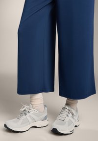 Svetlosivi športni čevlji z teksturiranim dizajnom in mrežnim detajlom, v kombinaciji z belimi rebrastimi nogavicami, oblečeni z širokimi mornarsko modrimi hlačami.