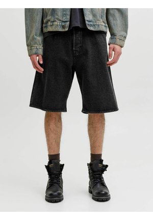 Osoba oblečená v ošúchaných čiernych riflových šortkách, čiernych šnurovacích topánkach s tmavými ponožkami a svetlom rifľovom saku, stojaca na jednoduchom pozadí.