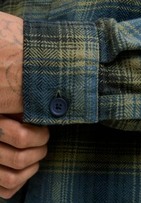 Blau-grünes karierter Hemd aus strukturiertem Stoff, mit einer geknöpften Manschette und einem dunkelblauen Knopf. Furchtbare Rautenmuster sichtbar.