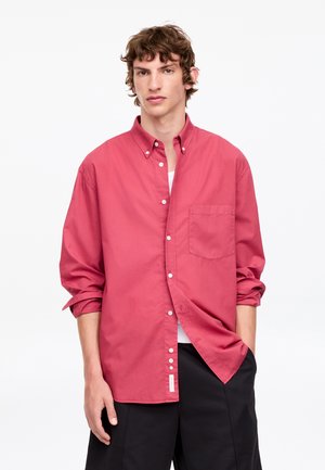 Jeune homme aux cheveux bruns ondulés portant une chemise rose ample boutonnée et un pantalon noir, debout devant un fond blanc uni.
