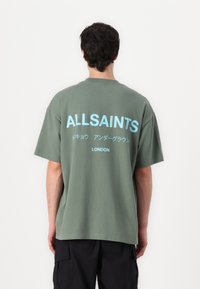 Zaļš pārspīlēts T-krekls no kokvilnas. Aizmugurē ir balti un gaiši zelti teksti, tostarp "ALLSAINTS" un "LONDON". Vienkāršs apaļā kakla dizains.
