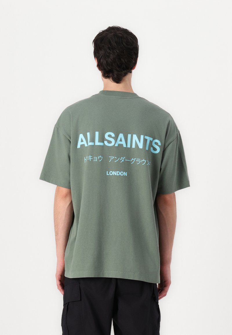 Zaļš pārspīlēts T-krekls no kokvilnas. Aizmugurē ir balti un gaiši zelti teksti, tostarp "ALLSAINTS" un "LONDON". Vienkāršs apaļā kakla dizains.