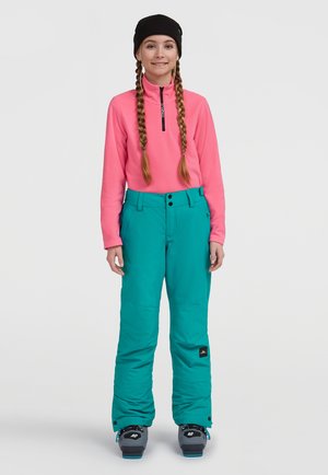 Kirkas vaaleanpunainen vetoketjullinen fleece-takki, teal-väriset lumihousut nappikiinnityksellä ja harmaat lumikengät sinisin yksityiskohdin. Yhdistetty mustaan pipoon.