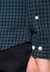 Groen en marineblauw geruit overhemd met knoopmanchetten, met witte knopen en een gladde textuur. Een zichtbare hand rust op een donkere stof.