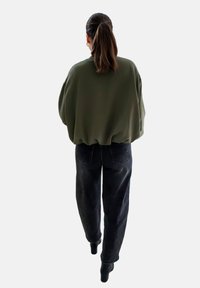 Overmaatse olijfgroene trui met verlaagde schouders, gecombineerd met donkere spijkerbroek en zwarte enkellaarsjes, gezien vanaf de achterkant tegen een neutrale achtergrond.