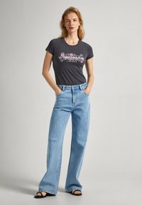 Camiseta gris oscuro con diseño gráfico floral y texto, combinada con jeans de campana azul claro. Sandalias negras completan el conjunto.