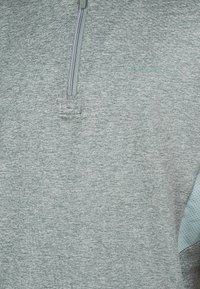 Hellgraues Kurzarmshirt mit einem Teilreißverschluss, glatter Textur und gestanzten blauen Akzenten an der Seite. Dezenter Glanz.