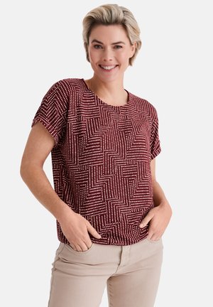 Lächelnde Frau mit kurzen blonden Haaren, die ein kastanienbraunes Kurzarmshirt mit weißen Punktemustern und beige Hosen trägt, die Hände in den Taschen.