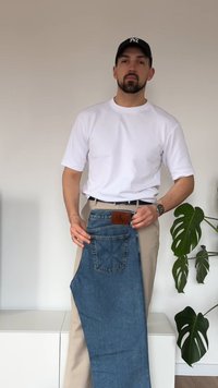 Μπλε τζιν με δερμάτινο σήμα, διαθέτουν τσέπες και διακριτικό σχέδιο ραφής, κρατούνται μπροστά από μπεζ chinos. Τα λευκό μπλουζάκι και το ρολόι φαίνονται.