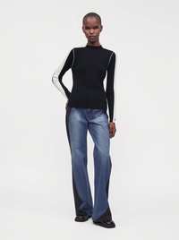 Femme portant un haut noir à manches longues avec des rayures blanches sur les manches et un jean large bicolore bleu et noir, debout sur un fond blanc.