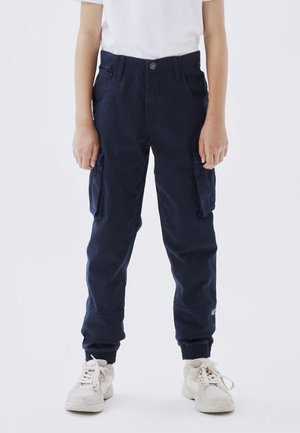 Enfant portant un pantalon cargo bleu marine avec des poches, des poignets élastiques, des baskets blanches et un t-shirt blanc uni à manches courtes, debout devant un fond blanc.