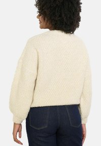 Krémovo sfarbený, textúrovaný pletený sveter s okrúhlym výstrihom a balónovými rukávmi, skombinovaný s tmavomodrými džínsami.