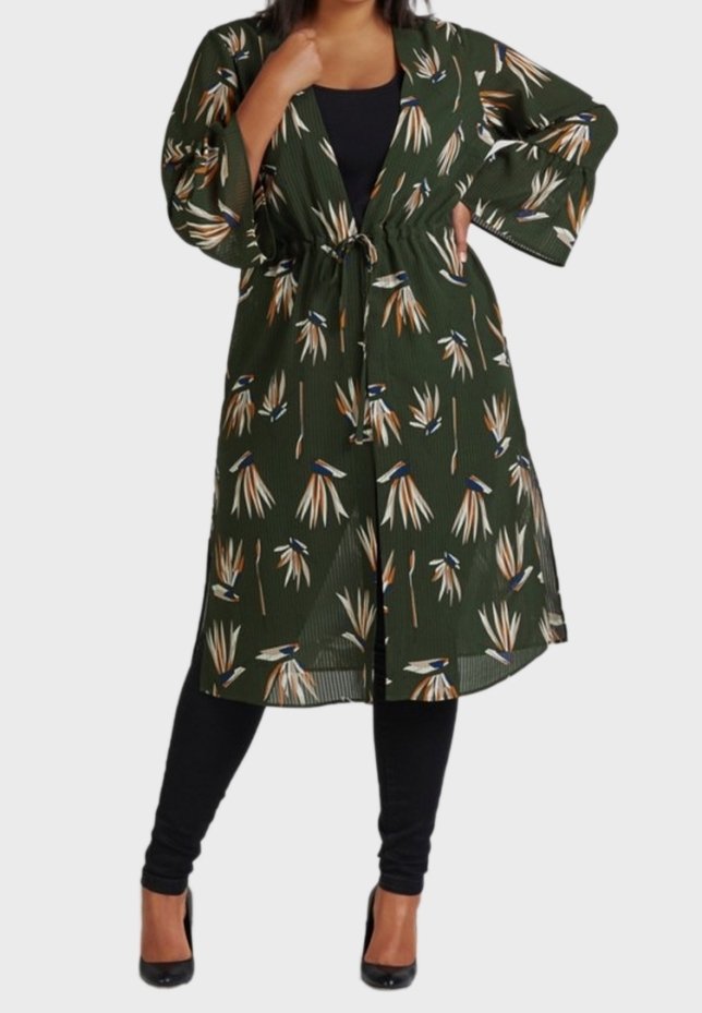 Femme portant une longue veste style kimono à fleurs vert foncé sur un haut noir, des leggings et des talons hauts noirs, debout avec une main sur la hanche.