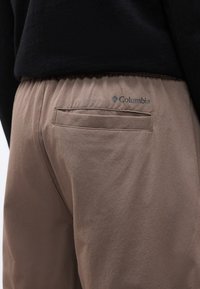 Κοντινό πλάνο στο πίσω μέρος παντελονιού ταουπ με ελαστική μέση και λογότυπο Columbia πάνω από μια οριζόντια πίσω τσέπη, φορεμένο με μαύρο τοπ.