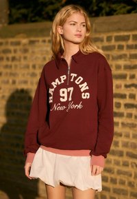 Hanorac cu fermoar de culoare burgundă, cu text alb "Hamptons 97 New York", asortat cu o fustă albă în straturi. Textură de material moale, croială lejeră.