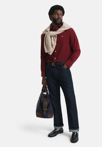 Rode shirt, beige trui over de schouders gedrapeerd, donkere denim jeans, zwarte schoenen, houdt een marineblauwe tas met bruin leren accenten vast.