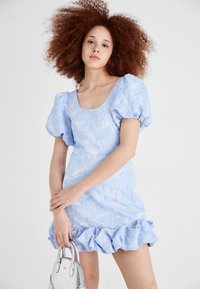 Light blue floral-patterned mini dress with puffed sleeves and a ruffled hem. Model holds a small silver handbag. Smooth texture.