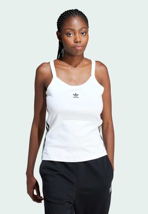 Top sin mangas blanco con escote redondeado, que presenta un pequeño logo negro de Adidas y franjas oscuras en los laterales, combinado con pantalones negros.